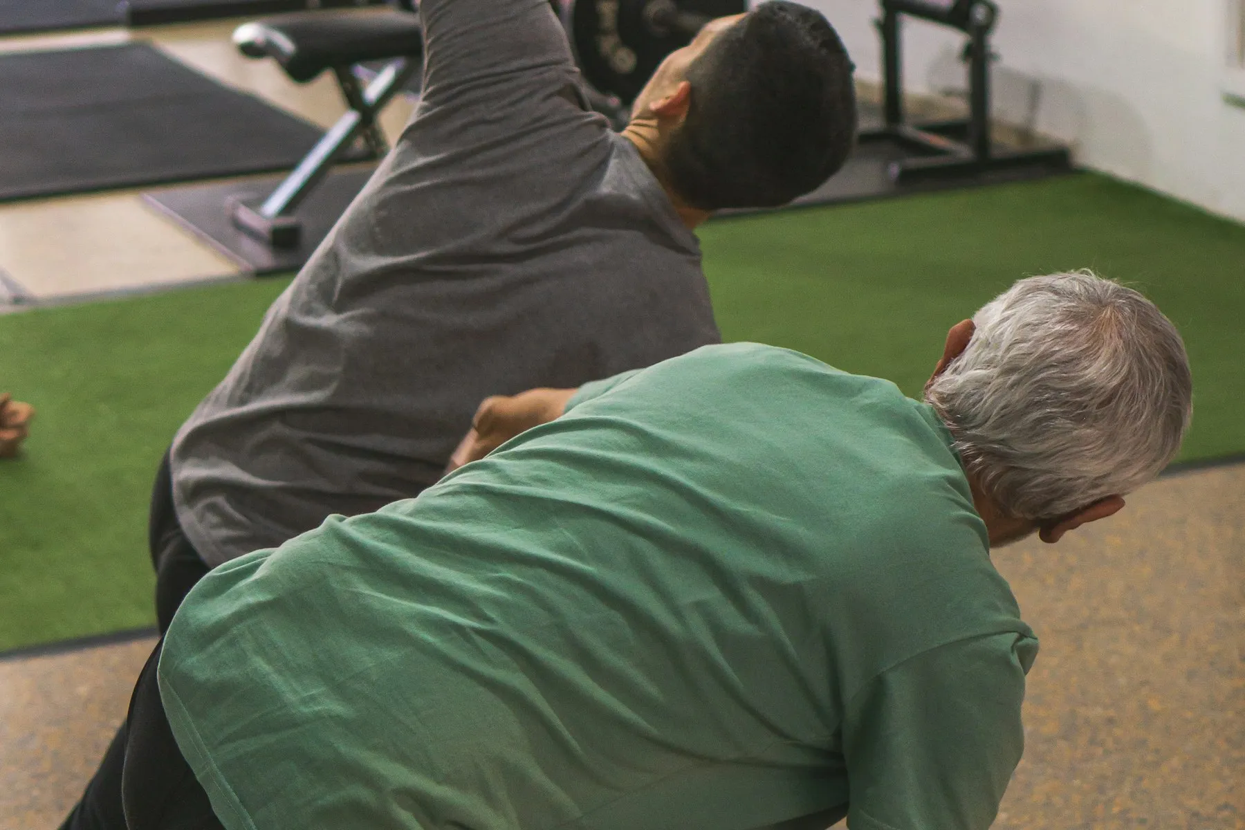 A white-haired man doing a stretching movement alongside an instructor in an exercise space
